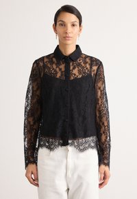 Juoda nėriniuota blusa su apykakle, mygtukų uždarymu ir ilgomis rankovėmis. Peršviečiamas nėrinių detalės prie pečių ir apatinėje dalyje su banguotais kraštais.