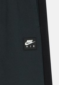 Tumša auduma panelis ar melnu Nike Air logo uzlīmi, izšūtu tuvu šuvju vietai divkrāsainā apģērba gabalā.