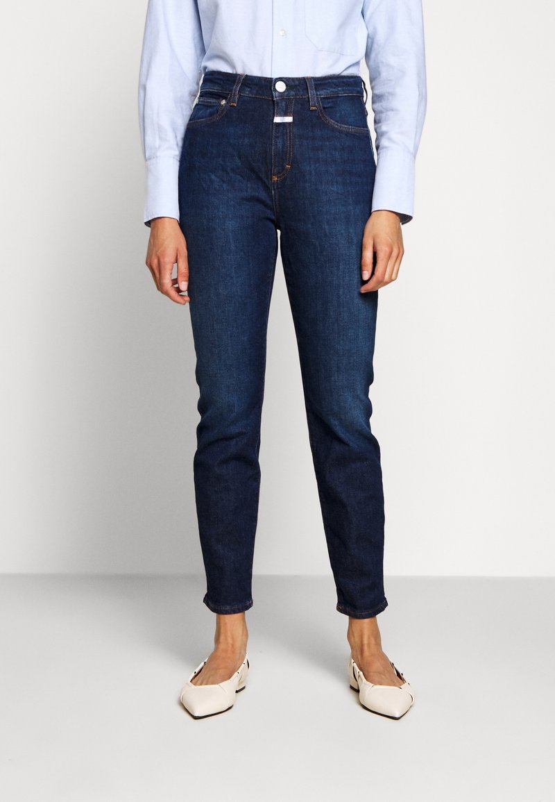 Jean bleu foncé taille haute, coupe droite, détails de couture subtils et fermeture par bouton à l'avant, associé à une chemise bleu clair.