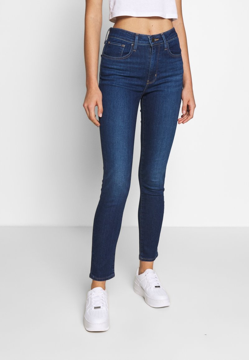 Jean skinny en denim bleu foncé, avec une taille haute, des poches avant, et une texture lisse, porté avec des baskets blanches.