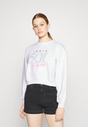 Sudadera corta blanca con el texto gráfico en gris y rosa "Levi's 501 Blues", combinada con pantalones cortos de tiro alto negros. Textura suave, corte relajado.