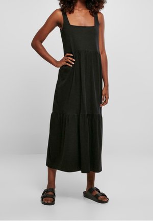 Femme portant une robe midi noire sans manches avec un décolleté carré et une jupe à volants, associée à des sandales noires, se tenant sur un fond gris.