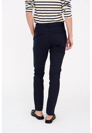 Person iført navy slim-fit bukser, sorte loafers og en hvid skjorte med sorte horisontale striber med lange ærmer, vist bagfra.