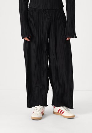 Black pleated wide-leg pants with a flowing design. Features an elastic waistband. Worn with white sneakers accented by red stripes.