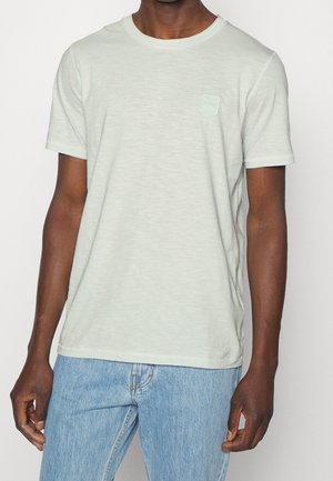 Man draagt een lichtgrijs T-shirt met korte mouwen en een klein logo op de borst, gecombineerd met lichtblauwe spijkerbroek, staand tegen een effen achtergrond.