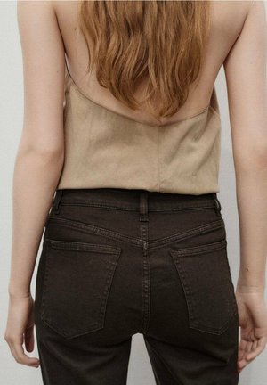 Femme vue de dos portant un haut beige sans manches et un jean taille haute foncé, avec de longs cheveux bouclés bruns tombant sur son dos.