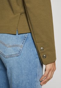 Pravá ruka u kyčle, viditelná kapsa na světle modrých džínách, oblékající olivově zelenou sako s dvěma knoflíky a detailem rozparku na zadní straně.