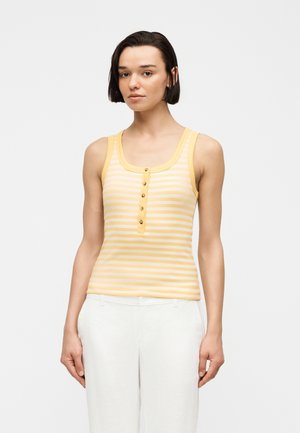 Νεαρή γυναίκα με κοντά σκούρα μαλλιά, φορώντας κίτρινη και λευκή ριγέ αμάνικη μπλούζα με κουμπιά και λευκό παντελόνι, στέκεται μπροστά σε απλό φόντο.