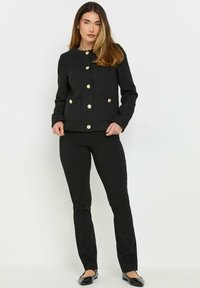 Mustaan tekstuurilla varustettu takki, jonka kullatut napit, pyöreä kaulus ja etutaskut, yhdistettynä mustiin housuihin ja kengättömiin kenkiin.