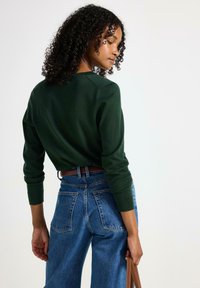 Grön stickad tröja med ribbade manschetter och rundad halsringning, parat med högmidjade blå denim byxor med vida ben, som har två bakfickor.