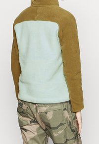 Zielona i brązowa polarowa kurtka z wysokim kołnierzem, z jasnobrązowym tylnym panelem i teksturowanym materiałem; zestawiona z spodniami w kamuflażu.