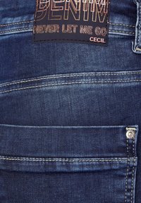 Vaqueros de mezclilla azul oscuro con costuras visibles, una etiqueta de tela rectangular que dice "DENIM NUNCA ME DEJARÁS CECIL" y un acento de botón.