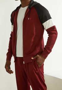 Chándal rojo con detalles en negro y blanco. Chaqueta con cremallera que incluye capucha, bolsillos laterales y tejido texturizado. Camiseta blanca debajo.