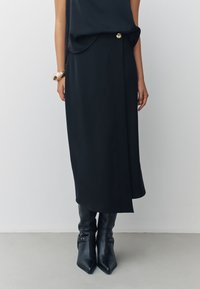 Gonna midi nera drappeggiata con spacco frontale e dettaglio di bottoni dorati, abbinata a stivali neri in pelle al ginocchio con fibbie decorative.