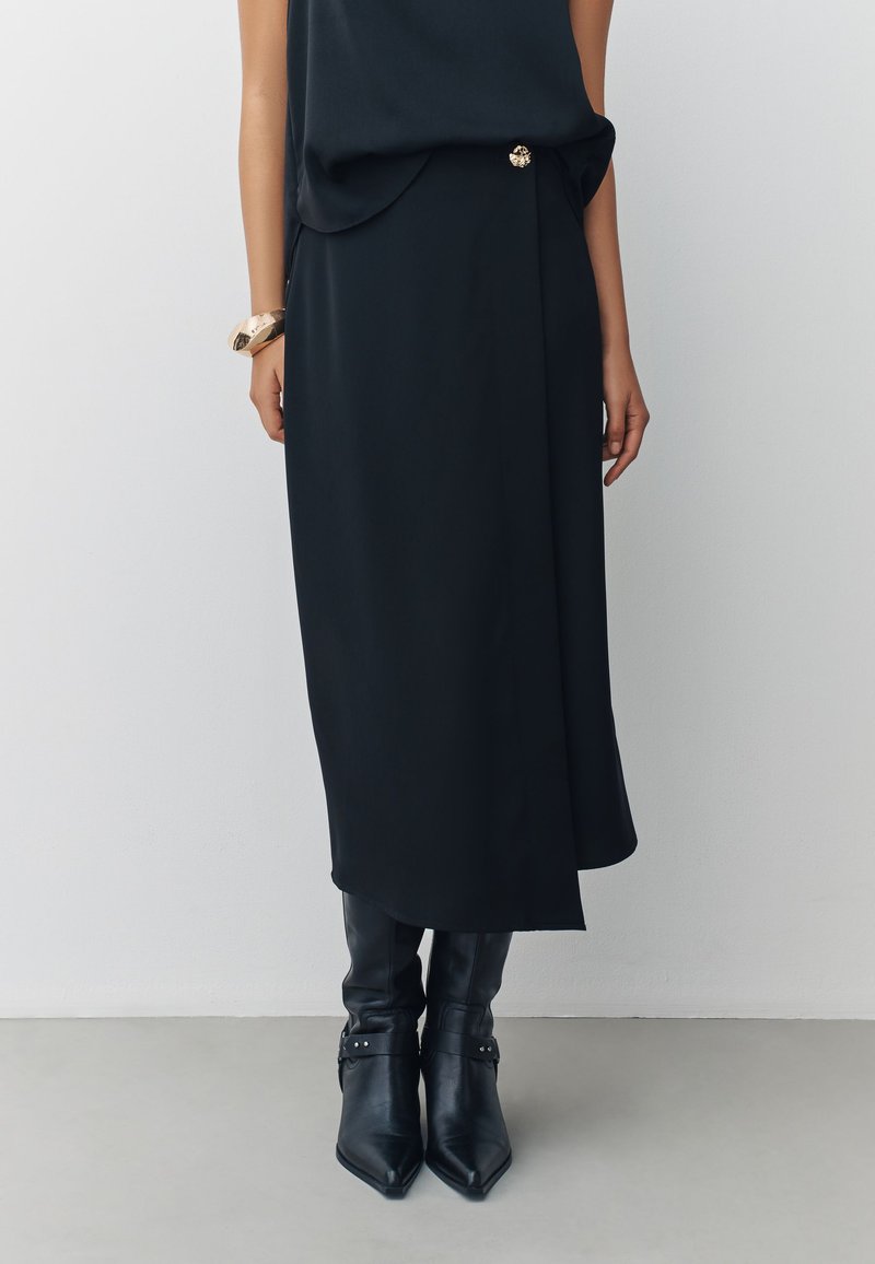 Gonna midi nera drappeggiata con spacco frontale e dettaglio di bottoni dorati, abbinata a stivali neri in pelle al ginocchio con fibbie decorative.