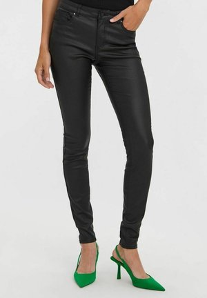 Czarne wąskie spodnie z ekologicznej skóry o gładkiej fakturze i pięcioma kieszeniami, zestawione z zielonymi, szpiczastymi szpilkami.