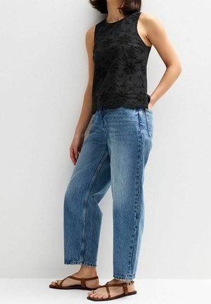 Femme portant un haut en dentelle florale noire sans manches, un jean bleu clair taille haute décontracté et des sandales plates marron, debout contre un mur blanc.