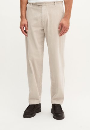 Beige Cordhosen getragen mit schwarzen Schuhen mit dicker Sohle, gezeigt an einem stehenden Mann vor weißem Hintergrund.