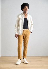 Witte overhemd met knopen over een zwarte tanktop, gecombineerd met mosterdkleurige corduroybroek en witte sneakers. Soepele texturen, casual pasvorm.