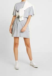 Grå bomullst-shirtklänning med avslappnad passform, med dragsko vid midjan och en vit sweatshirt över axlarna. Vita högstövlar.