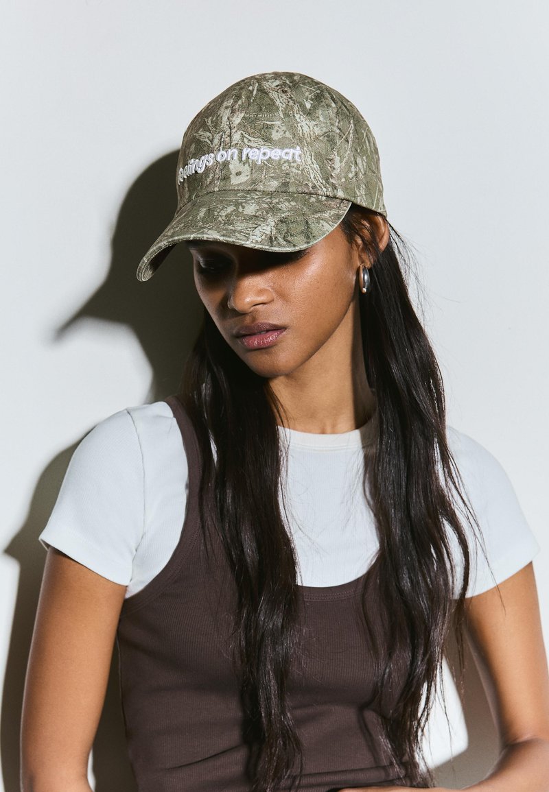 Femme portant une casquette de baseball camouflage avec le texte « Settings on repeat », un t-shirt blanc à manches courtes et un débardeur marron, regardant vers le bas.