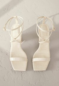 Weiße Sandalen mit krokodilprägung, offenen Riemen und quadratischem Absatz, die mehrere dünne Riemen aufweisen und über eine glatte Textur sowie ein minimalistisches Design verfügen.