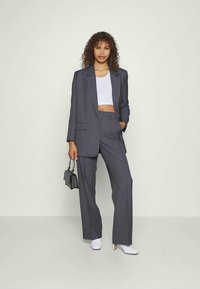 Mulher em pé vestindo um fato cinza oversized com um top branco curto, botins brancos até o tornozelo, segurando uma pequena bolsa preta contra um fundo simples.