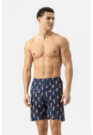 Hemdloser Mann mit lockigem Haar, der dunkelblaue Shorts mit einem bunten Vintage-Pin-up-Mädchen-Print trägt und vor einem schlichten weißen Hintergrund steht.