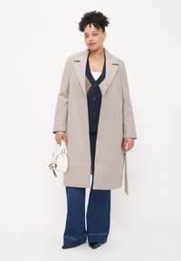 Cappotto di lana beige con un design sartoriale, colletto ampio e taglio dritto. Abbinato a un cardigan blu navy e jeans in denim svasati, con una borsa bianca in mano.