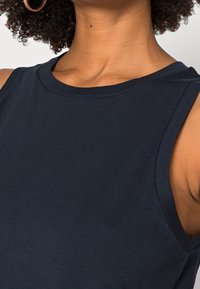 Kvinna iklädd en ärmlös marinblå topp och stora guldörhängen, visar hals och axlar med lockigt hår synligt.