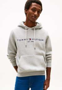 Joven con sudadera con capucha gris claro de Tommy Hilfiger con logo y vaqueros azules, de pie con las manos detrás de la espalda sobre un fondo blanco.