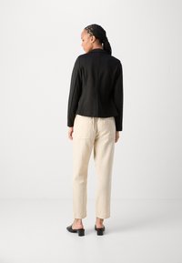 Svart, skräddarsydd blazer med slagkrage, kombinerad med ljusbeige, texturerade byxor. Modellen bär svarta klackmules.