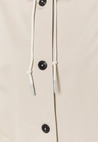 Beige kledingstuk van stof met zwarte knopen en witte trekkoorden, met een gestructureerd materiaal en een ontwerp dat de functionele sluitingsdetails benadrukt.