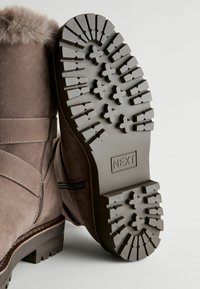 Brown suede ankle boot with a fur cuff, featuring a textured brown rubber sole with cross-patterned treads and a visible brand logo.