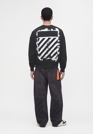 Homme portant un sweat-shirt noir avec des rayures graphiques blanches et un jean noir à jambes larges avec une étiquette orange, debout de dos sur un fond uni.