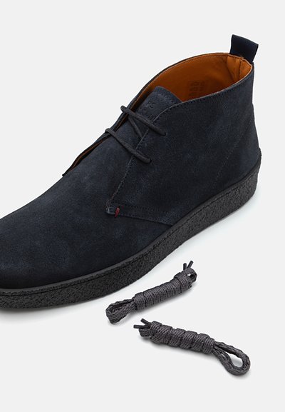 Zapato chukka de ante negro con una suela negra texturizada, con cordones grises al lado. El interior tiene un forro naranja y costuras mínimas.