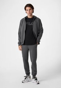 Survêtement athlétique gris avec veste zippée, T-shirt noir avec logo "FILA" et pantalon fuselé, complétés par des baskets blanches.
