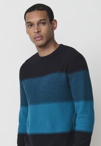 Adīta džemperis ar apaļu kaklu, kas apveltīts ar zilu un melnu horizontālu joslu rakstu. Strukturēts augšējais josls un gludas apakšējās daļas.