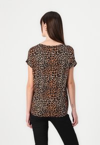 Femme aux longs cheveux bruns portant un haut à manches courtes imprimé léopard et un pantalon noir, de dos sur un fond clair uni.