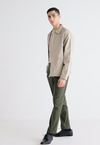 Veste légère beige avec fermeture éclair et poches latérales, associée à un pantalon vert foncé et des chaussures noires. Design simple, texture lisse.