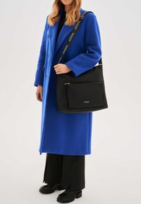 Bolso negro de hombro con un bolsillo frontal con cremallera, con un material texturizado y una correa con marca. Lleva puesto un abrigo azul y pantalones negros.