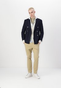 Blazer doppiopetto blu navy, camicia crema a bottoni con motivo floreale sottostante, pantaloni beige e sneakers bianchi. Texture lisce evidenti.