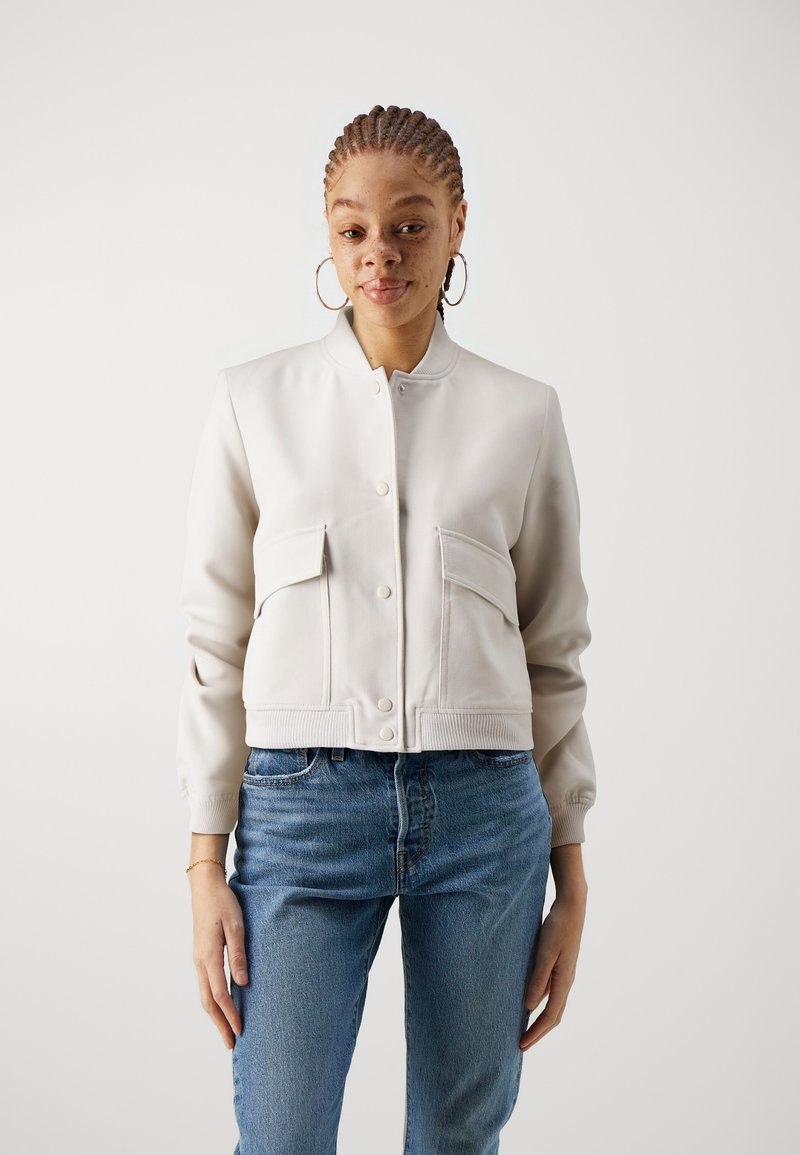 Veste bomber cropped beige clair avec deux poches à l'avant, fermetures à pression et ourlet côtelé, associée à un jean bleu. Texture lisse.