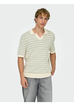 Mladić svijetlosmeđe kose u krem i bež prugastom pletenom polo majicom kratkih rukava i širokim sivim trapericama, stoji na neutralnoj pozadini.