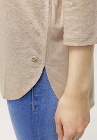 Top beige texturizzato con orlo arrotondato, con un piccolo bottone decorativo di lato, abbinato a jeans blu.