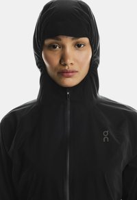 Must kollane veekindel jakk kõrge kaelusega ja kapuutsiga, millel on eeslt tõmblukk ja logo vasakul küljel. Sile, mati tekstuur.