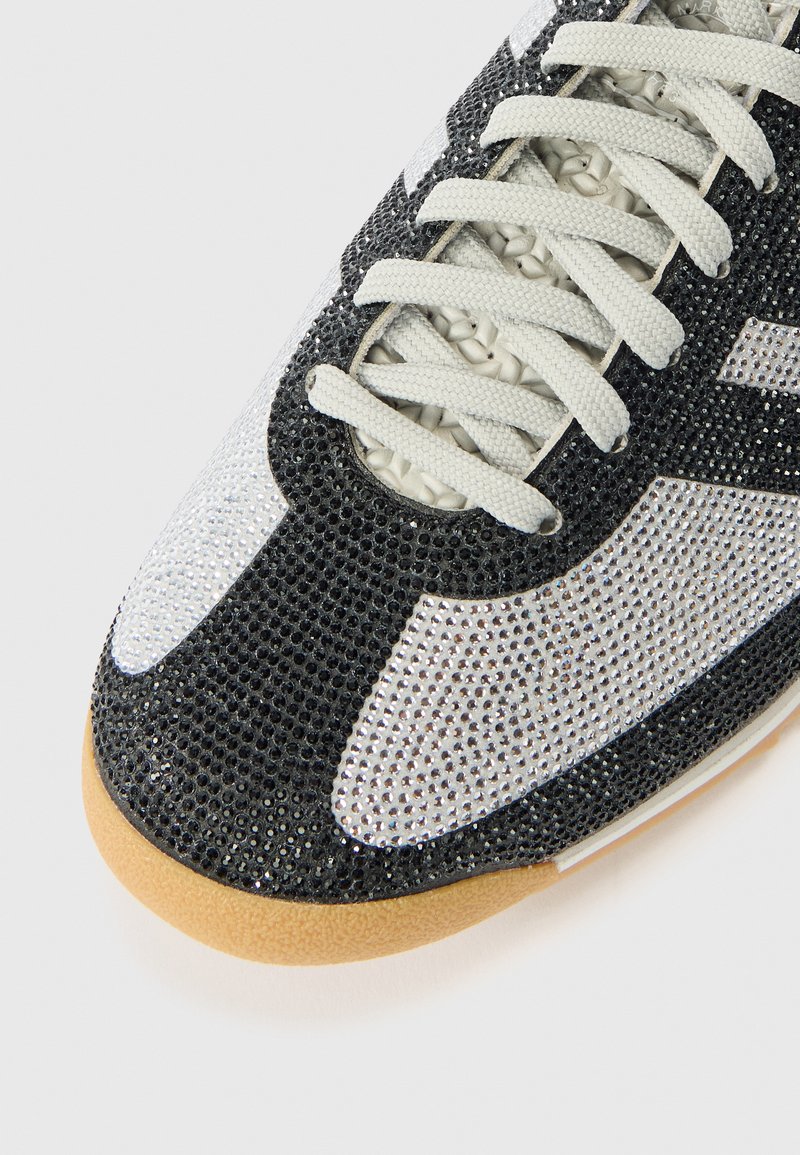Sneaker con tomaia in strass neri e argento, con puntale bianco e suola in gomma beige. I lacci sono grigio chiaro.