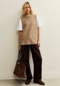 Tankleurige gebreide mouwloze top over een wit T-shirt, gecombineerd met donkerbruine wijde broekspijpen en een bruine boodschappentas. Platte bruine instapschoenen.