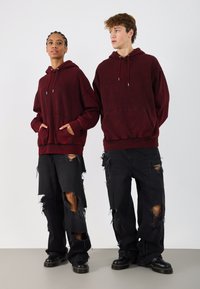 Burgundy kapucnis pulcsik elülső zsebekkel, viselnek kopott, fekete nadrágot, melyeken nagy lyukak találhatóak, és fekete csizmát. Egyszerű háttér.