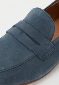 Kék velúr loafer, sima textúrával, kiemelkedő övpánttal és varrott részletezéssel a széleken. Bélelése világosbarna.